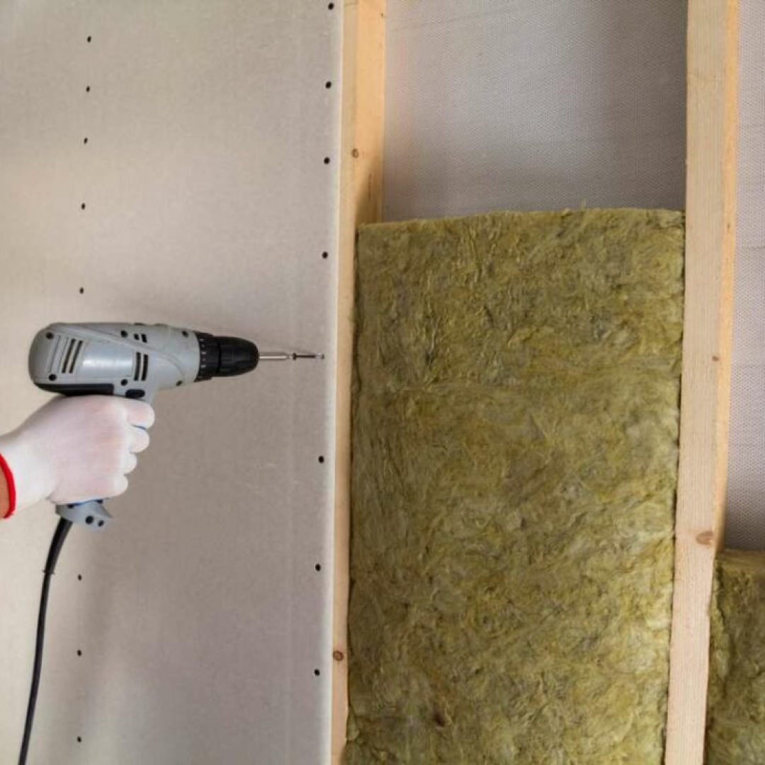 Close-up of worker hand with electric screwdriver fastening drywall to wooden timber frame with insulation mineral rock wool staff. Warm home, low energy bills, construction and renovation concept.