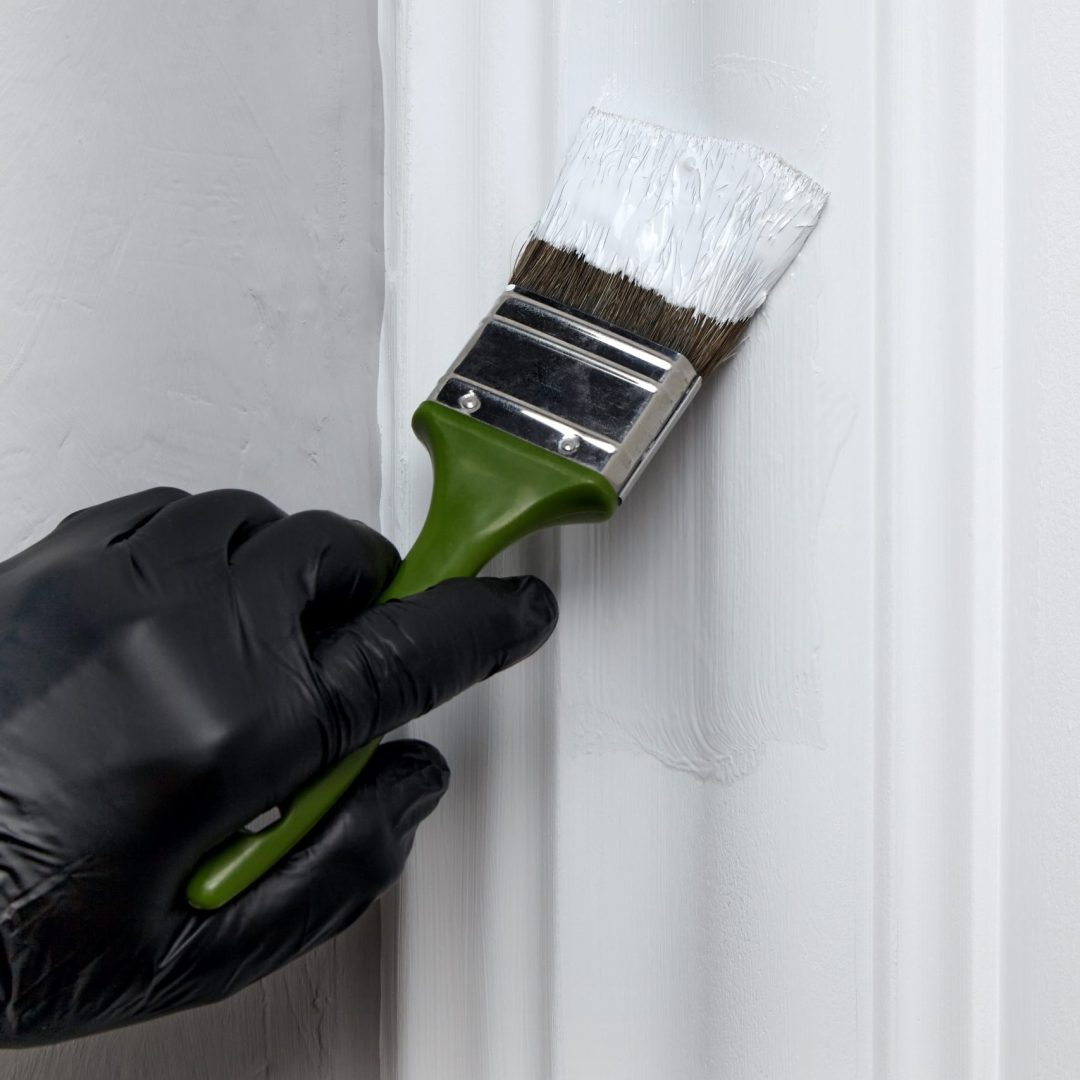 A hand in a black rubber glove paints a decorative door frame with white paint with a brush. Repair and restoration of the apartment with your own hands
