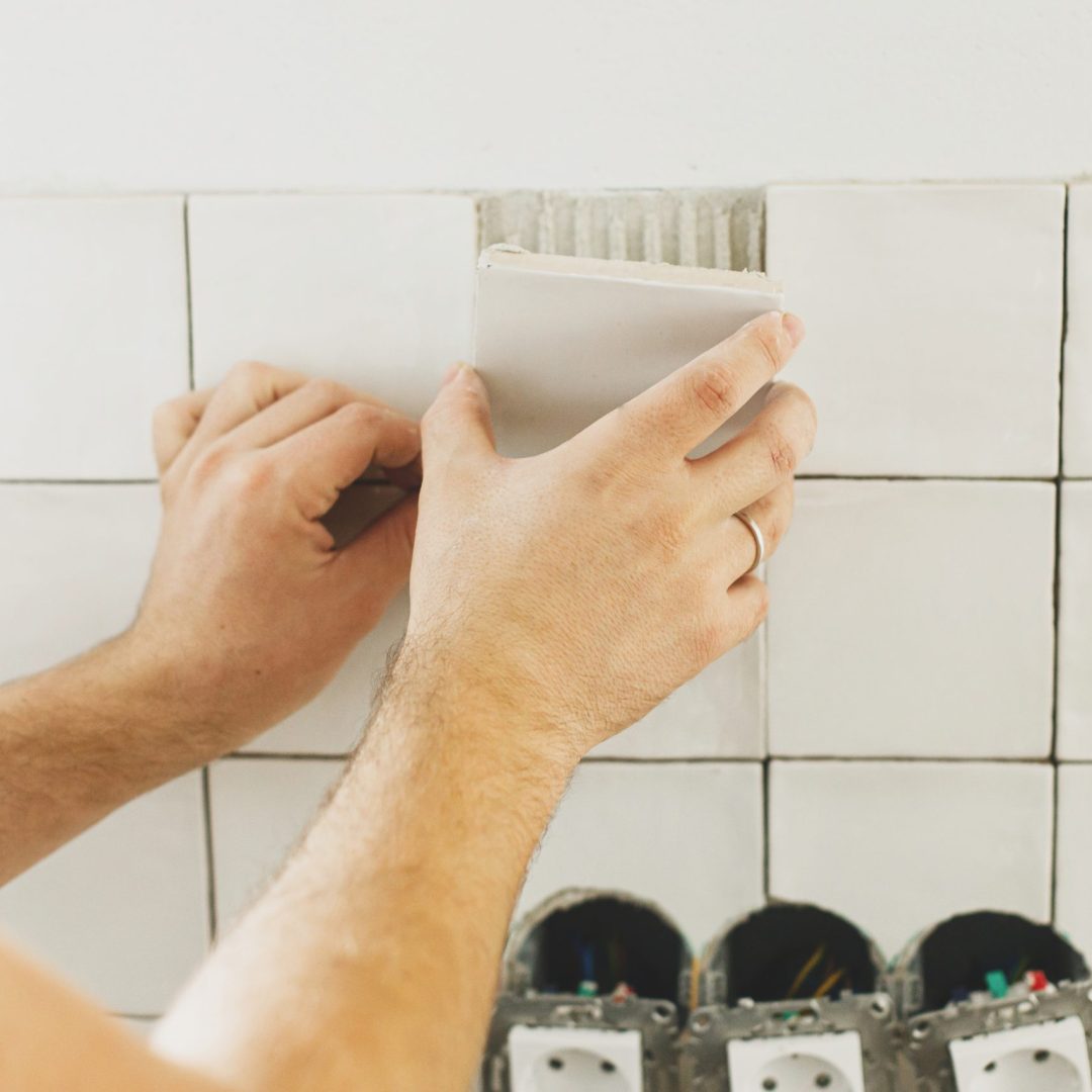 Hands laying modern square tile on adhesive close up. Worker installing stylish white tiles on plaster wall. Renovation